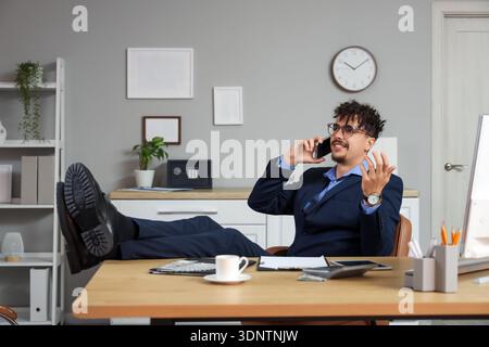 Un giovane direttore di banca con le gambe sul tavolo che parla al telefono nel suo spazio di lavoro in ufficio Foto Stock