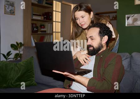 Giovane donna adulta caucasica e uomo di mezza età caucasico seduto sul divano con un laptop insieme, donna appoggiata sulla spalla dell'uomo che guarda lo schermo, entrambi concentrati sul dispositivo Foto Stock