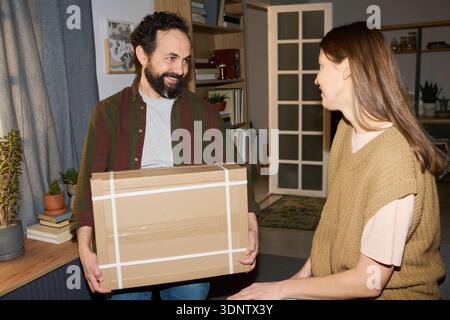Uomo di mezza età caucasico che tiene in mano una scatola di cartone mentre sorride alla donna di mezza età caucasica, entrambi interagiscono in ambiente domestico con scaffali e piante visibili sullo sfondo Foto Stock