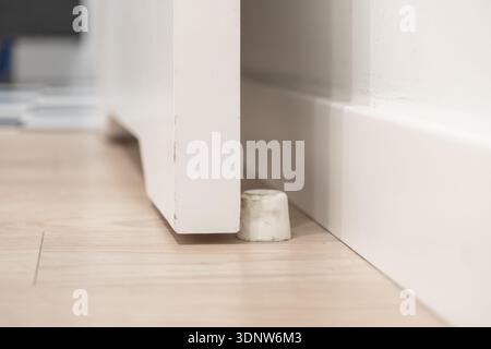 Vista ravvicinata di un piccolo fermaporta bianco cilindrico collocato su un pavimento di legno chiaro, che mantiene una porta bianca minimalista in modo sicuro e socchiusa in un'area residenziale Foto Stock