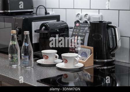 Cracovia, Polonia - 30 novembre 2025: Bollitore elettrico nero, macchina per il caffè, due tazze bianche e acqua in bottiglia esposta con cura in una cucina elegante Foto Stock
