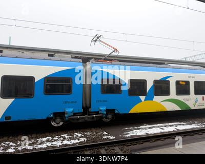 Poznan, Polonia - 30 novembre 2025: Vista laterale del treno POLREGIO, con livrea blu, bianca, gialla, verde, con pantografo, parcheggiato su binari in mezzo al wi Foto Stock