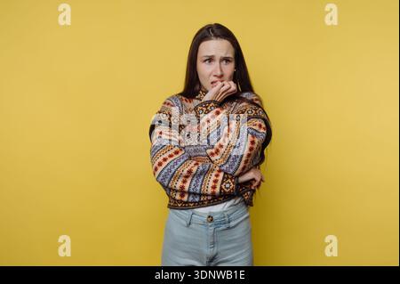 Una giovane donna sta con un'espressione preoccupata, la sua mano che copre la bocca. Indossando un maglione colorato, appare nel profondo del pensiero, surrou Foto Stock