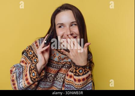 Vestita con un comodo maglione a motivi geometrici, una giovane donna sorride e tiene il telefono all'orecchio. La sua espressione giocosa rivela che è più affilata Foto Stock