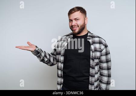 Un giovane allegro sorridente si presenta con sicurezza con un sorriso ampio, vestito con una camicia a quadri e una maglietta nera. Si comporta in modo invitante come se accogliesse qualcuno Foto Stock