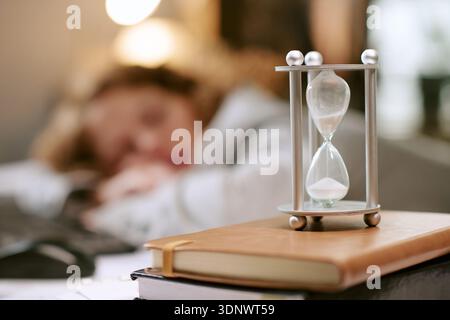 Giovane donna adulta che dorme alla scrivania con clessidra in primo piano Foto Stock