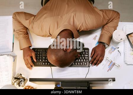 Uomo nero che appoggia la testa sulla tastiera mentre lavora in ufficio Foto Stock