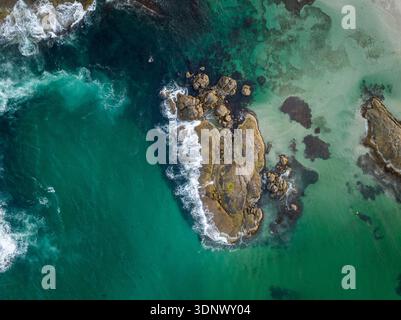 Una scena costiera sopraelevata mostra le onde che si infrangono su una barriera corallina rocciosa circondata da profonde acque blu e turchesi con motivi subacquei visibili. Foto Stock