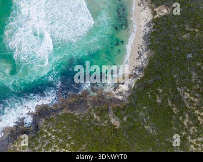 Prospettiva aerea dall'alto verso il basso che mostra le onde turchesi che si infrangono su una costa rocciosa, incontrando una piccola insenatura sabbiosa accanto alla fitta vegetazione costiera. Foto Stock