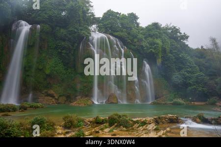 Un'ampia cascata scorre su rocce coperte di muschio in una tranquilla piscina circondata da una fitta vegetazione tropicale all'interno del complesso delle cascate Ban Gioc. Foto Stock