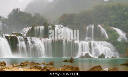 La cascata Ban Gioc scorre in ampie tende a gradoni sulle scogliere calcaree in una tranquilla piscina turchese, con rocce e sponde sabbiose in primo piano. Foto Stock