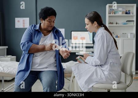 Donna nera anziana che applica gel sul braccio dimostrando il problema della pelle al medico femminile Foto Stock