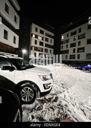 Notte nevosa in un complesso residenziale con auto parcheggiate e un luminoso lampione stradale. Foto Stock