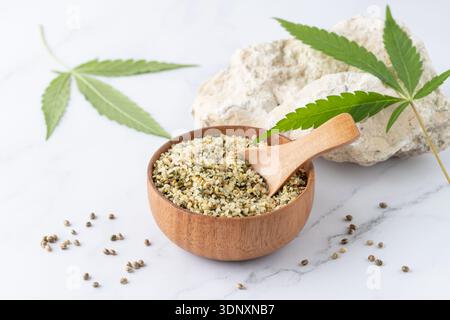 Semi di canapa schiacciati sgusciati in ciotola di legno come ingrediente per mangiare e foglie di cannabis su sfondo di marmo bianco. Semi di cannabis come supercibo, morbidi Foto Stock