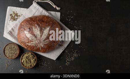 La pagnotta di farro appena sfornata con semi di canapa tritati è decorata con foglie di cannabis fatte di farina su sacchetto di tessuto di canapa su dorso grunge marrone Foto Stock