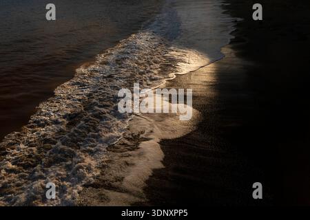 Le onde dolci scivolano sulle sabbie scure di una spiaggia delle Asturie in Spagna, con la luce del sole dorata che si riflette elegantemente sulla superficie delle acque, creando una S Foto Stock