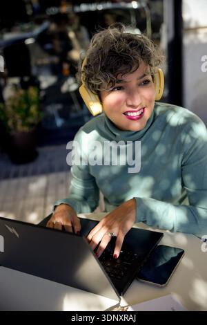 Donna adulta felice che si diverte a lavorare su un notebook all'aperto, indossando le cuffie. La luce solare filtra creando un motivo di luce e ombra sulla sua f Foto Stock