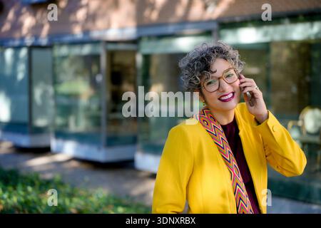 Donna d'affari sorridente in una giacca gialla vibrante, che parla al telefono fuori. La donna adulta, sicura di sé e esperta di tecnologia, ama lavorare all'aperto Foto Stock