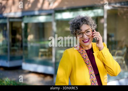 Donna d'affari adulta felice esperta di tecnologia all'aperto al telefono, che trasuda fiducia e successo. Indossando occhiali da vista e una giacca gialla vibrante, lei è l'emblema Foto Stock