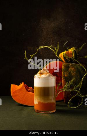 Un bicchiere di latte di zucca e spezie condito con panna montata si staglia su uno sfondo scuro, circondato da zucche rustiche e viti decorative Foto Stock
