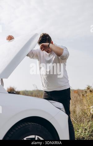 Un giovane si trova accanto alla sua auto elettrica bianca, ispezionandola sotto un cielo limpido. È vestito casualmente e sembra concentrato sulla comprensione della sua veh Foto Stock