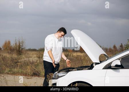 Un giovane uomo si trova di fronte alla sua auto elettrica bianca con il cofano aperto e sembra perplesso. Il veicolo è parcheggiato su una strada di campagna sotto una nuvola Foto Stock