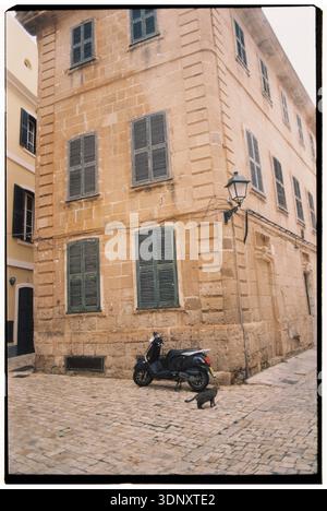 Una strada pittoresca di Minorca presenta un vecchio edificio in pietra con persiane verdi, uno scooter nero parcheggiato e un gatto vagante su pavimentazione di ciottoli. Foto Stock