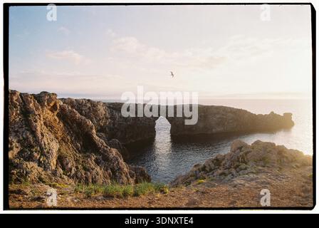 Una splendida vista di un arco roccioso naturale su un mare calmo al tramonto a Minorca. La scena cattura la tranquillità della costa mediterranea, con Foto Stock