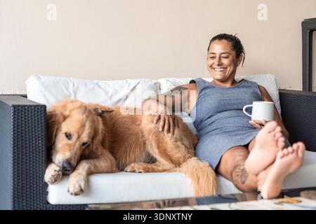 Una donna d'affari ama rilassarsi a casa con il suo cane, seduto su un divano con una tazza calda in mano. Sorride contentosamente, prediligendo la torta Foto Stock