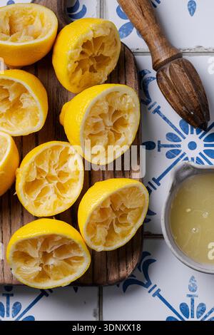 Vista dall'alto di diversi limoni spremuti a metà su un asse di legno con una ciotola in ceramica di succo di limone. Un alesatore di legno si trova nelle vicinanze su una pacca bianca e blu Foto Stock