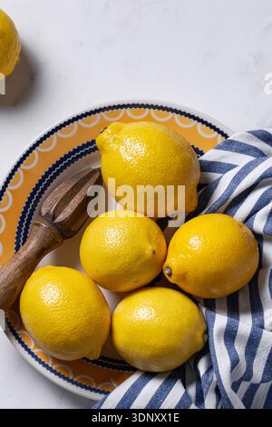 Vista dall'alto dei vivaci limoni gialli su un piatto colorato con una fresa di legno. Un panno a strisce blu e bianco aggiunge una texture, il tutto su una marba bianca Foto Stock