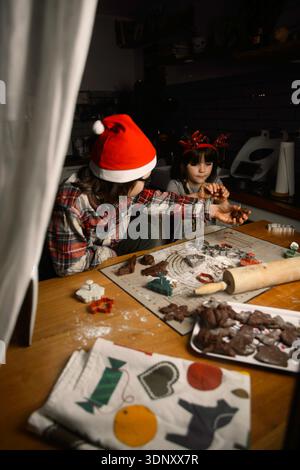 Due sorelle trascorrono un pomeriggio festoso preparando biscotti natalizi in una cucina illuminata. Indossando accessori a tema natalizio, creano insieme Foto Stock