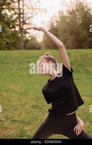 Una giovane donna pratica yoga in un tranquillo ambiente all'aperto, circondato da lussureggianti prati verdi e alberi. Tiene con grazia una posa yoga, incarnando tran Foto Stock