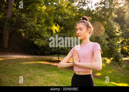 Una giovane donna pratica yoga all'aperto, circondata da una vegetazione lussureggiante. Sta con calma con le mani in una posizione di preghiera, abbracciando la tranquillità in un Foto Stock