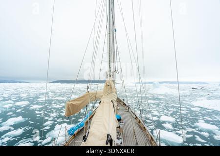 Una barca a vela naviga nelle acque ghiacciate della Groenlandia, circondata da blocchi di ghiaccio galleggianti e montagne lontane, catturando la serena ma dura bellezza di Th Foto Stock