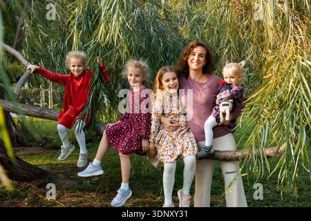 Una madre gioiosa e le sue figlie si divertono all'aperto, appollaiate su un albero caduto. I bambini, indossando abiti colorati, irradiano felicità e tog Foto Stock
