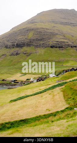Pittoresca vista di un villaggio collinare sull'isola di Vagar, le Isole Faroe, con case tradizionali annidate tra lussureggianti campi verdi e la suggestiva rugge Foto Stock