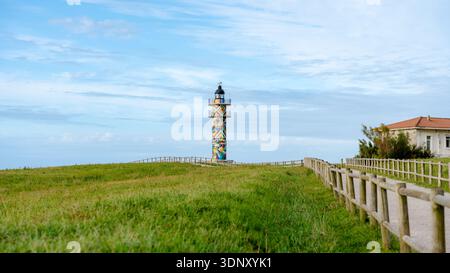 Il colorato Faro di Ajo si erge maestosamente contro un cielo blu limpido, circondato da prati verdi lussureggianti ed edifici tradizionali in Cantabria, Nor Foto Stock
