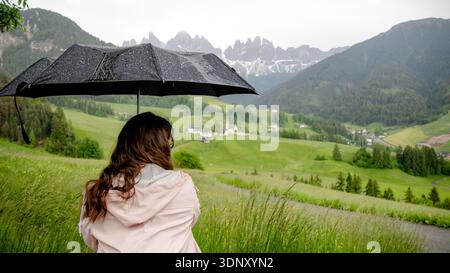 Una donna sotto l'ombrello gode della vista sulla Chiesa di Santa Maddalena in Val di Funes, con le maestose montagne della catena delle Odle sullo sfondo c Foto Stock