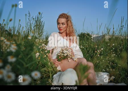 Un'immagine serena di una madre in attesa in un prato illuminato dal sole, trasuda amore e tranquillità mentre siede tra le margherite, simboleggiando nuovi inizi e natura Foto Stock