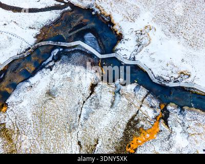 Vista aerea di un fiume serpeggiante che attraversa un paesaggio innevato e strutturato con una passerella in legno che attraversa le sue acque scure, le sorgenti termali e Hverage Foto Stock