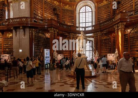 Austria, Vienna, 6 agosto 2025: Turisti che visitano la Biblioteca Nazionale austriaca con la statua centrale e le gallerie di libri. Foto Stock