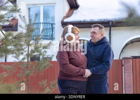 Una coppia di mezza età amorevole in piedi insieme in inverno vicino a casa di famiglia. Felice coppia anziana che si abbraccia fuori casa loro. Invecchiamento sano, per tutta la vita Foto Stock