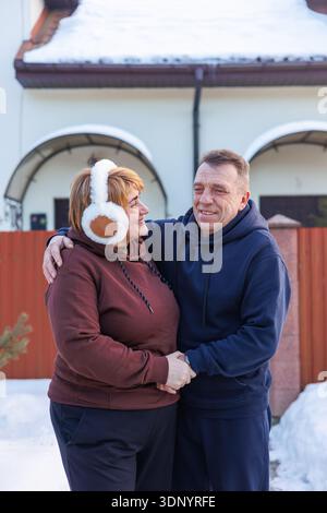 Una coppia di mezza età amorevole in piedi insieme in inverno vicino a casa di famiglia. Felice coppia anziana che si abbraccia fuori casa loro. Invecchiamento sano, per tutta la vita Foto Stock
