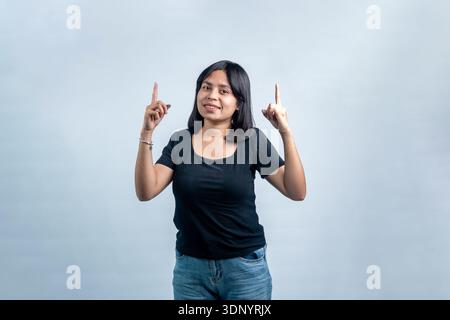 Giovane donna del sud-est asiatico con una camicia nera che guarda la macchina fotografica e punta verso l'alto. Ritratto in studio isolato su sfondo grigio chiaro. Foto Stock