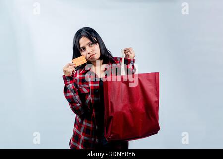 Una giovane donna del sud-est asiatico con una camicia di flanella rossa che sembra triste e sexy, mentre tiene in mano una carta di credito e una borsa della spesa su uno sfondo bianco. Foto Stock