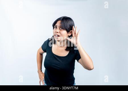 Ritratto di una giovane donna del sud-est asiatico che si piega in avanti con la mano a orecchio, che sembra curiosa e sorpresa mentre ascolta su uno sfondo bianco. Foto Stock