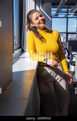 Donna negli anni '30 appoggiata al davanzale illuminato dal sole in ufficio, con top senape e smartwatch Foto Stock