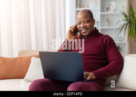 Uomo nero maturo che parla al telefono mentre lavora al computer portatile a casa Foto Stock
