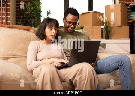 Partner diversi e felici che si rilassano sul divano nella nuova casa, guardano film sul laptop e sorridono. Giovane coppia multietnica che naviga online sui siti web di mobili, godendo di una pausa nel giorno del trasloco. Foto Stock